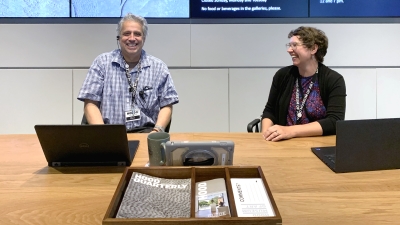 Two adults sit behind a long wooden desk. They are greeting museum visitors as they enter an atrium space. They are smiling and laughing. Behind them are screens with information written in white text on a black background.