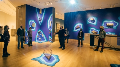 A group of museum visitors stand in a gallery with dark blue walls and ceramic art hanging on them. The ceramic art looks like fishing traps and nets. A central figure is speaking and leading the tour.