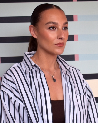 A portrait photograph of a woman sitting in three-quarter profile. She has tan skin and her dark hair is pulled back into a low ponytail. She is wearing a white and black oversized button-down shirt and a black shirt underneath.