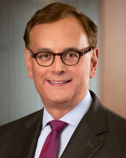 A headshot of a middle-aged caucasian man with brown hair and glasses, he is wearing a suit and tie.