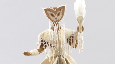 A figure made of tanned hide, wearing a mask, and holding a fan with feathers sits stop a woven basket.