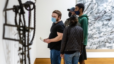 College-age students explore a museum exhibition of contemporary art that focuses on the idea of lines. There is a metal sculpture slightly out of focus in the foreground and behind the students is a large abstract painting.