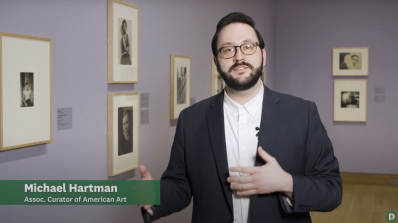 A still from a video of a adult male standing in a museum gallery with lavender walls discussing an installation of black and white photographs of old Hollywood.