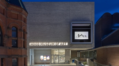 A photograph of the Hood Museum of Art's north facade and entrance taken after sunset. There is a window on the facade displaying a black and white video work.
