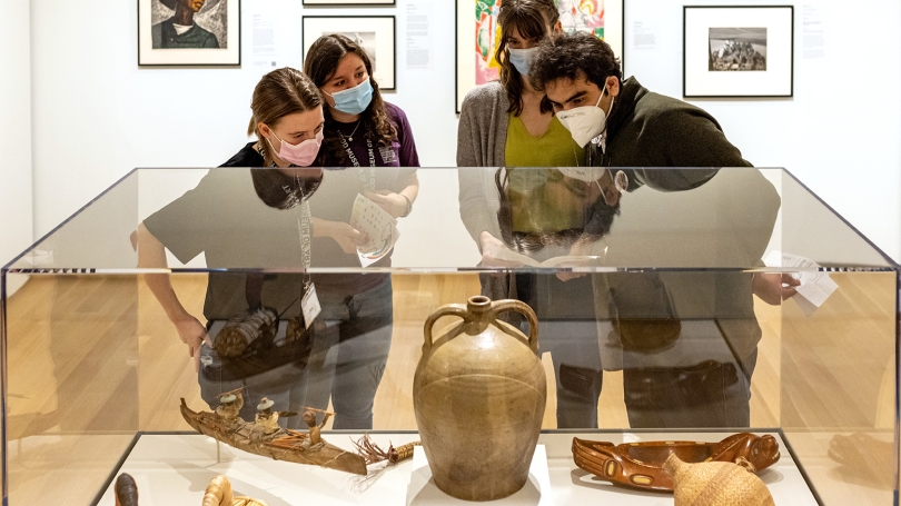 A small group of 4 college-aged students stand around a case of objects in a museum gallery. The people are wearing masks and looking at Native American and American objects.