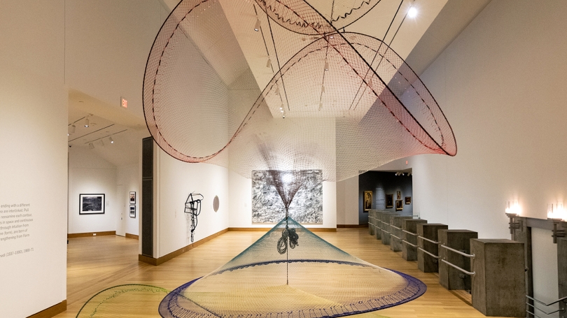 Suspended sculpture made of hand-dyed fishnets with four distinct sections that are connected together. The top two section mirror each other in shape while the bottom two are congruent. 