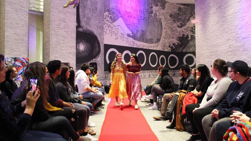 At the center of the photo, two laughing women in a yellow dress (left) and a pink dress (right) stroll down a light red carpet. On either side of the photo, college students sit in rows watching them walk.