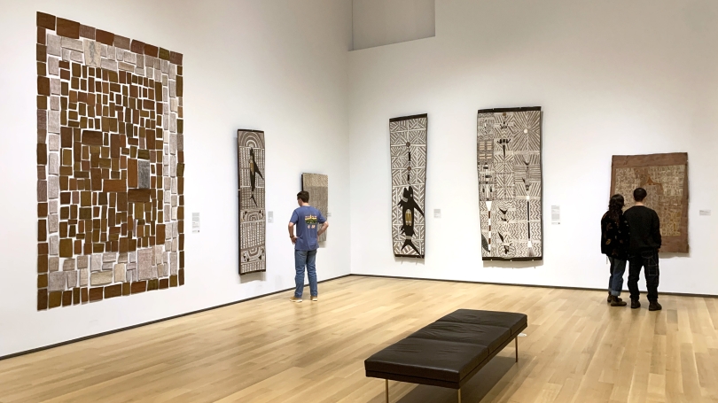Three college-aged students stand in a gallery with high ceilings. The walls are white and on them are Aboriginal Australian bark paintings.