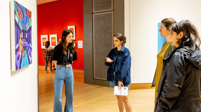 One person stands infront of a painting and discusses its meaning with three onlookers