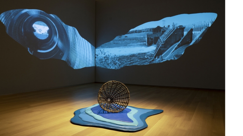 A photograph of a museum installation that features a two-channel video in the shape of whale's fins. In the foreground there is a fishing basket trap made of ceramics and sitting on a wooden platform painted in different shades of blue.
