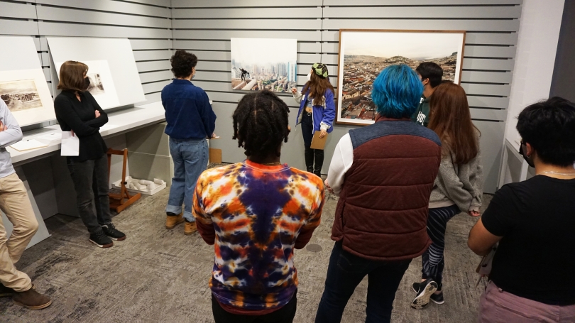 A group of college students stand in a classroom, all are wearing masks. They are circle around and watching a fellow student give a presentation about a photograph that hangs on the wall behind them