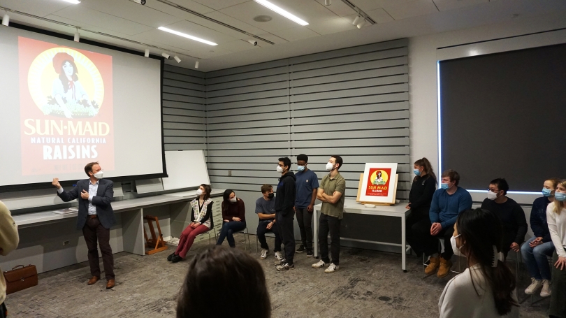 A class of college students stand in a smart classroom, surrounded by various works on paper, and are looking at the front of the classroom to their professor who is discussing one of the works projected onto a large screen.