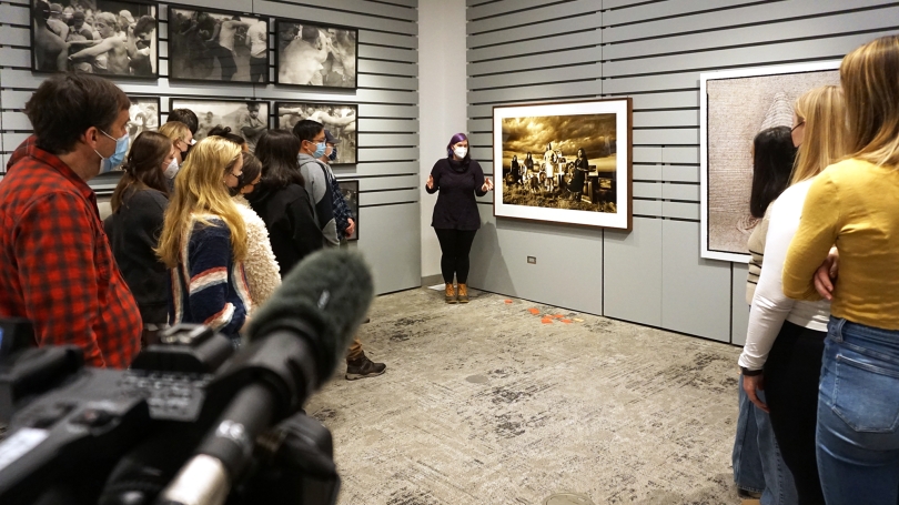 A group of medically masked college students form a semi-circle around a woman who is discussing or teaching the students about the photographs that hang on the wall behind her.