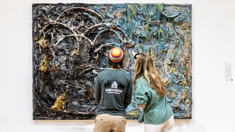 Two college-aged students stand in a museum gallery with white walls and with their backs to the camera. They are looking at and discussing a large assemblage work in front of them. The work of art is made out of found objects and cloth.