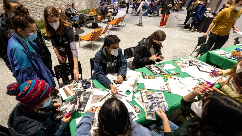 College-aged students sit around a table, that is covered in a green tablecloth, and create collages using various paper materials.