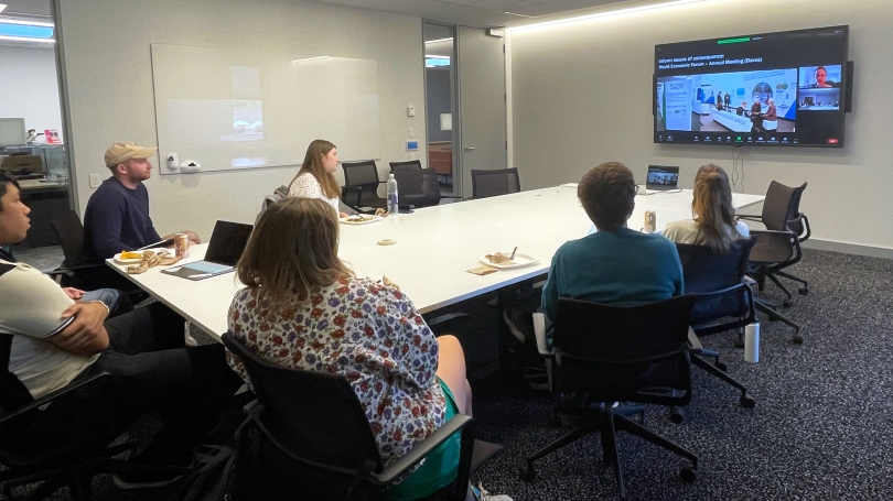 A group of college students sit around a long white table and are looking at a screen at the far end of the table. Someone is on screen presenting to the group.