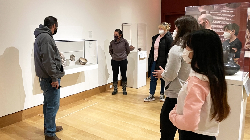 A group of medically masked college students stand in a gallery in a semi-circle formation. The gallery they are standing in is installed with Native American art, mostly ceramics and photography. Two artists are presenting to the group of students.