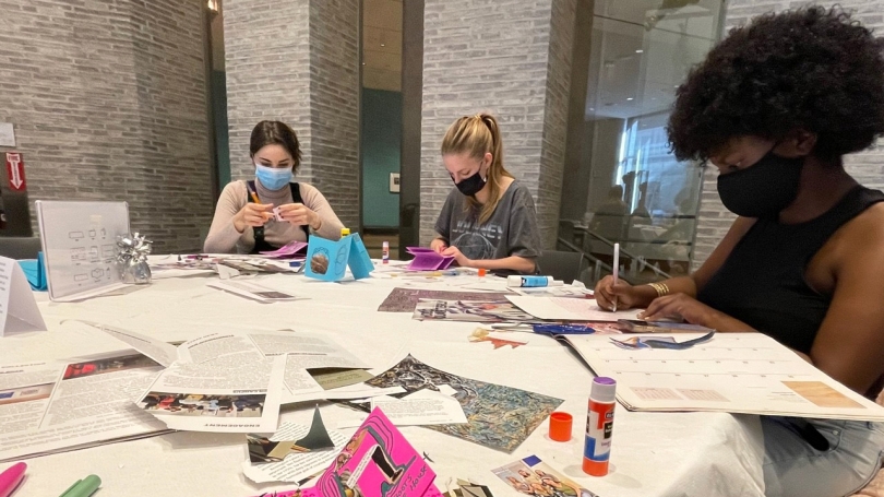 Three college-aged females sit at a round table crafting. They are cutting out materials from magazines and newspapers.