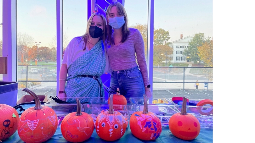 Two medically masked college-aged females stand behind a table adorned with a white table cloth. On the table in front of them is a line of decorated orange pumpkins.