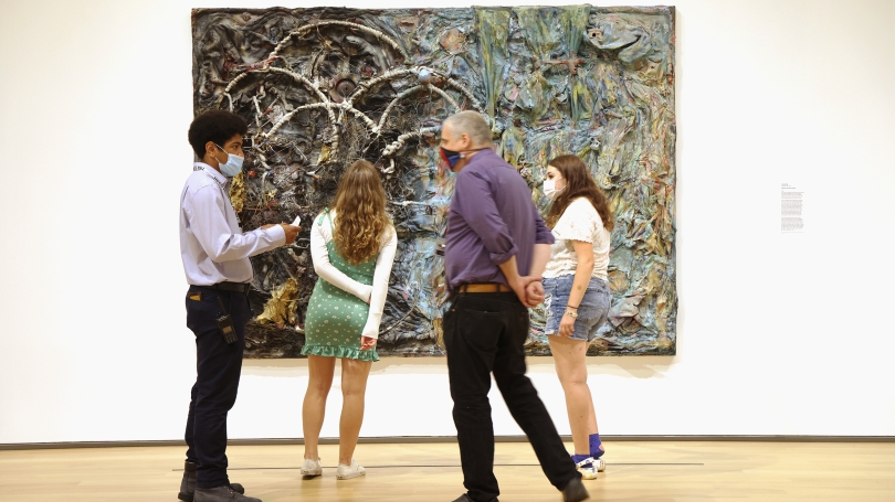 In the background, two college-aged people are looking closely at a large assemblage work of art that is created with found objects, paint, and cloth. In the foreground two male individuals are medically masked and talking with one another.