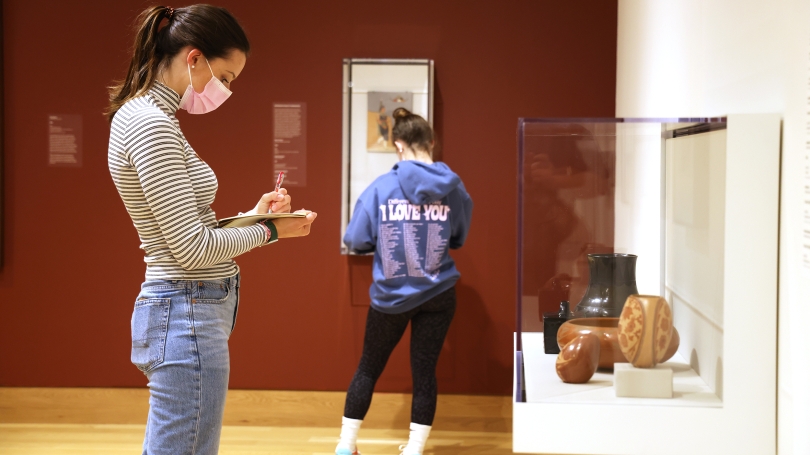 A high school student is looking at a sculpture inside of a case. She is holding a pad of paper and a pencil and making notes. She is wearing a striped turtle neck and jeans and is shown in profile.