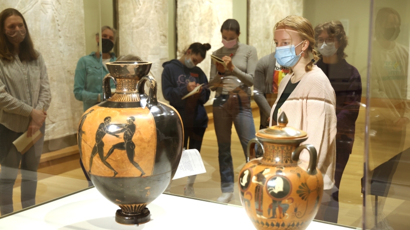 A group of high school students stand in a gallery. In the foreground is a girl with a blue medical mask on. She is looking at an ancient Greek Amphora inside of the museum floor case.