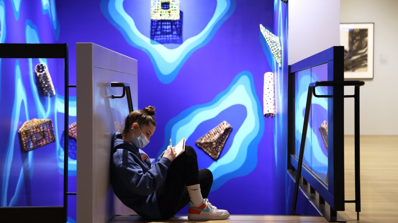 A high school student, wearing a blue medical face mask, sits at the top of a flight of stairs in a museum gallery that is painted dark blue. She is writing on a pad of paper with a pencil. Works of ceramic art hang on the walls around her.