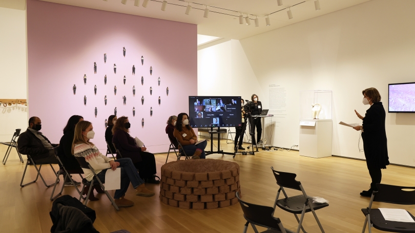 Adults sit on black stools in a museum gallery that is installed with contemporary ceramic art made by Native American artists. The adults are watching a presentation.