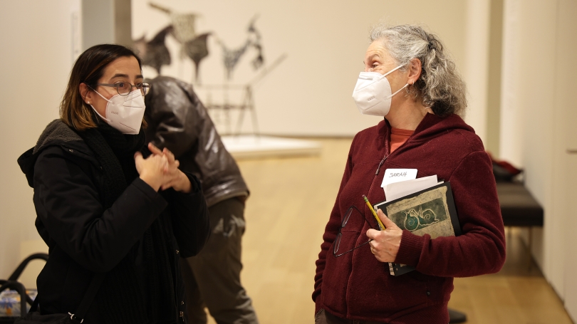 Two adult women, wearing white N95 medical masks, are laughing and talking inside of a museum gallery.