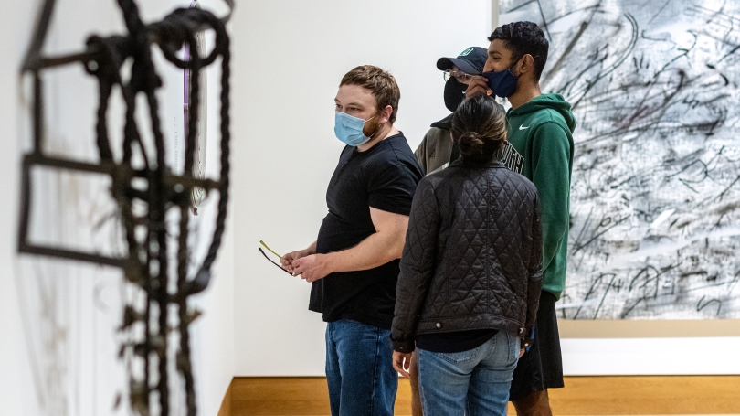 College-age students explore a museum exhibition of contemporary art that focuses on the idea of lines. There is a metal sculpture slightly out of focus in the foreground and behind the students is a large abstract painting.
