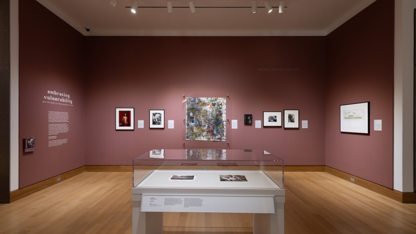 a view of Embracing Vulnerability: Gay Intimacy in the Context of AIDS installed in Gutman Gallery. The walls are a reddish brown and there is art hanging on them.