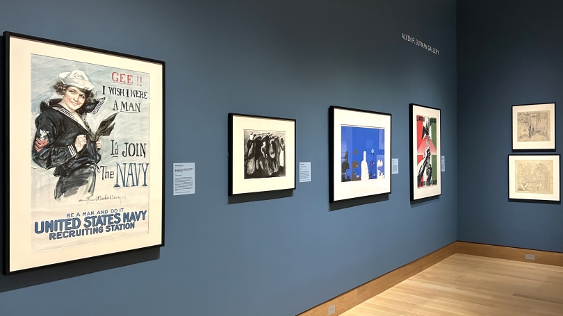 A museum gallery installation mostly comprised of World War one and two posters. The walls are painted a dark blue.