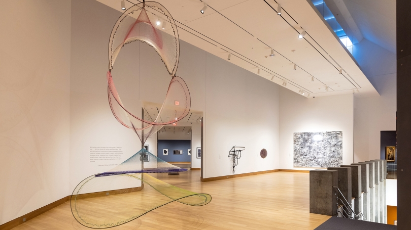 A photograph of a museum gallery and installation of contemporary art, including a large kinetic sculpture made of colorful netting in the foreground.