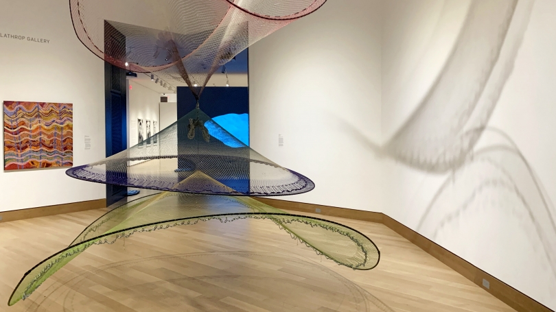 A photograph of an exhibition installation which includes a an Australian Aboriginal painting and a large netted sculpture that hangs from the ceiling. 