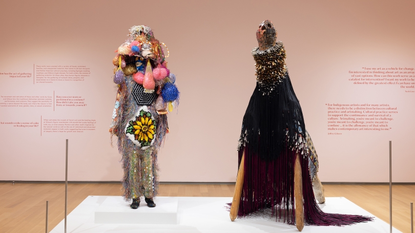 Two larger-than-life figural sculptures stand in the middle of a room painted pink. Both sculptures are made out of found or recycled materials.