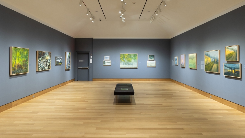 A museum gallery with dark blue-gray walls, installed with contemporary and realistic landscape paintings.