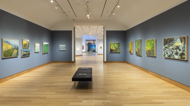 A museum gallery with dark blue-gray walls, installed with contemporary and realistic landscape paintings. The photographs shows the next gallery with features works by the same artist.