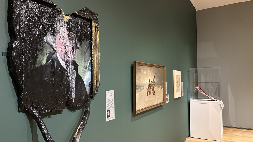 A museum gallery installation with gray and forest green walls. In the foreground of the installation image is a work of art that looks like a portrait of President Washington in a book that has been burnt.