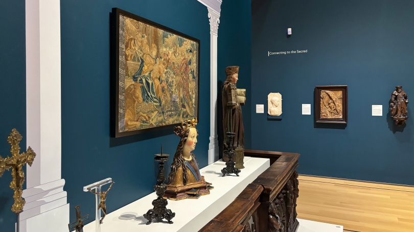 At the center of a room with blue walls sits a large wooden table with a glass case containing multiple sculptures of various sizes, shapes and materials. Behind it, hangs a large colorful tapestry.