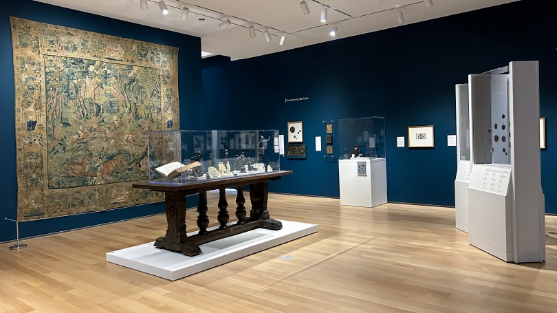 At the center of a room with blue walls sits a large wooden table with a glass case containing multiple sculptures of various sizes, shapes and materials. Behind it, hangs a large colorful tapestry. 