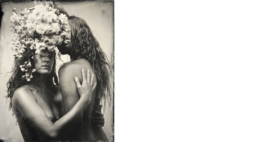 Two nude biological female individuals embrace in a hug. There are flowers in their hair. One individual's face is turned toward the camera, but is obscured by the flowers, and the other is facing away from the camera. The image is black and white.