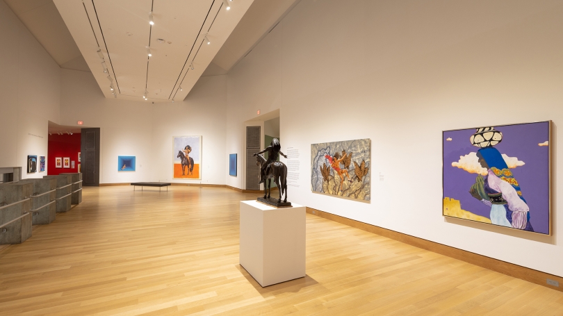 A museum gallery installed with paintings and one sculpture in the center of the installation. The gallery walls are painted white and the ceiling is very high. 