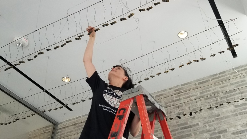 A woman stands on a ladder and hangs small sound devices from the cieling.