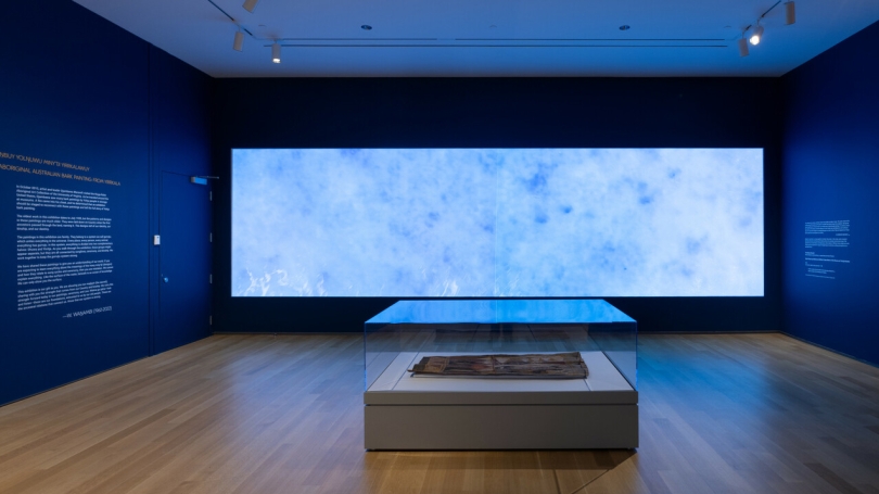 A museum gallery with the walls painted a deep dark blue. There is a case on the floor in the middle of the room which holds the oldest known Indigenous Australian bark painting. In the background is a large two screen video projection.