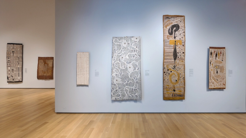 An installation of Indigenous Australian bark paintings hang in a museum gallery with a light gray wall.
