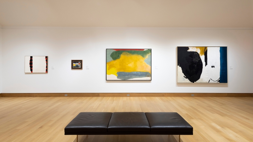 A museum gallery with white walls with three large abstract paintings with fields of color, and one smaller work. There is a bench in the foreground of the image.