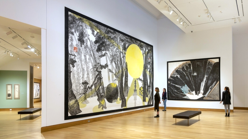A group of three young women stand in front of monumental works on paper that almost reach floor to ceiling in a gallery with a cathedral height ceiling. The works are by a contemporary Korean artist using the Korean ink and brush painting technique.