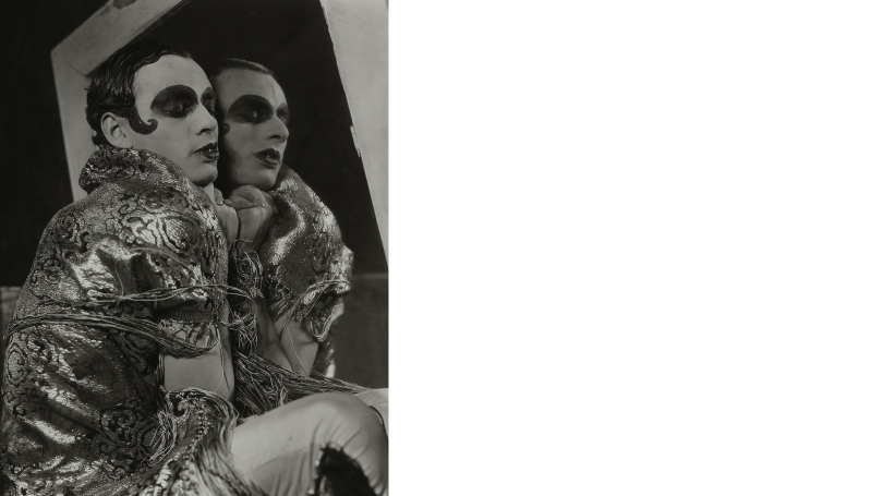 A black and white photograph of a drag queen. They are in profile and leaning up against a mirror so that you can see the other side of their face in reflection. They are wearing a metallic looking jacket and heavy dark eye makeup.