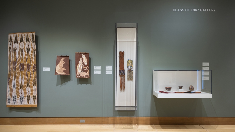 A museum gallery with grayed blue-green walls displays works of art that employ ochre. There are two Indigenous Australian bark paintings hanging on the wall as well as two wall cases displaying works of art.