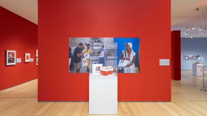 A museum gallery with red walls and high ceilings. There is a case with beach balls against a wall with three large vinyl photographs of older Cuban women depicted. Inside the case the beach balls and one bag say I love, symbolized by a red heart, Cuba.